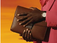 Person wearing a pink suit holding a brown textured leather clutch with a watch on their left wrist against an orange background.