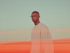 Portrait of a man wearing glasses and a light sweater standing against a blurred background with orange and green hues.