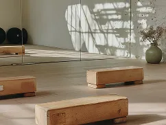 Wooden Pilates blocks arranged on a sunlit wooden floor in a minimalist exercise studio with a mirror and a vase of dried flowers.