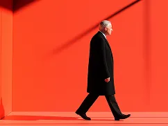 Man in a black coat walking inside a brightly lit orange room with sharp shadows.