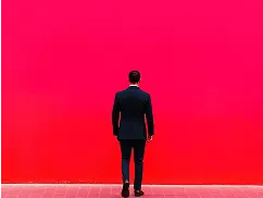 Man in a black suit standing and facing a bright red wall.