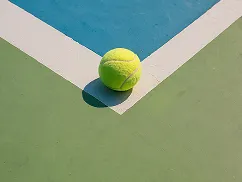 Tennis ball resting on the corner of a tennis court with blue and green sections divided by a white line.