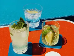 Three drinks on a red and blue table: a mojito with mint, a glass of water with lemon, and a cocktail with lime wedges.