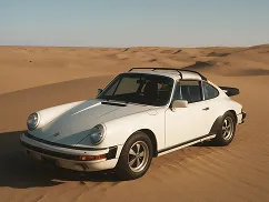 White classic Porsche 911 parked on desert sand dunes under a clear blue sky.