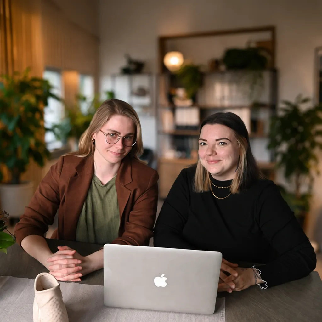 Die Femvestory-Gründerinnen Lena und Elisabeth sitzen lächelnd an einem modernen Arbeitsplatz mit Laptop – Unabhängige Finanzplanung für Frauen auf Augenhöhe.
