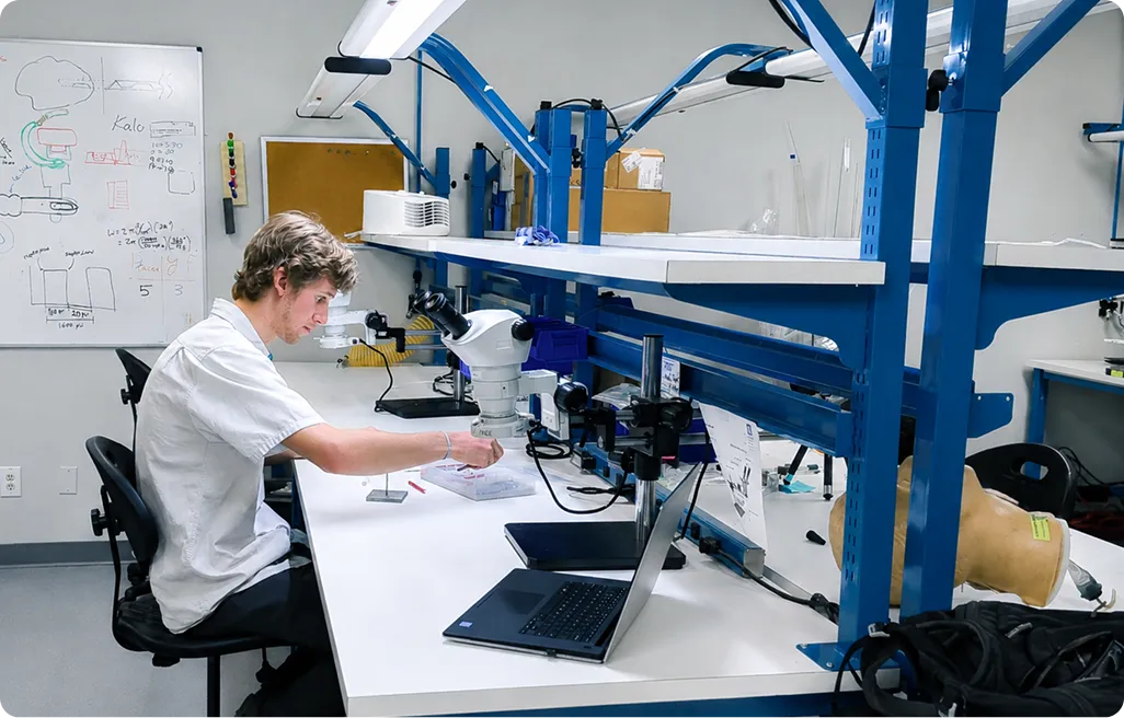 Person working with microscope and plastic containers at a lab bench with laptop and whiteboard nearby.