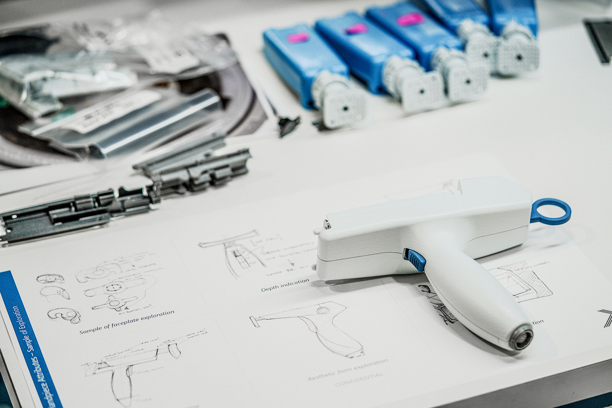 White pistol-grip medical handpiece prototype resting on a printed page of design sketches, with mechanical components and package parts in background