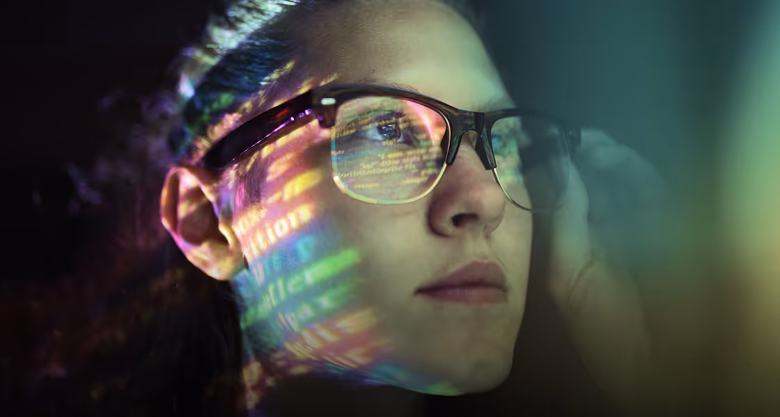 Close-up of a woman wearing glasses with colorful code reflections on her face and lens, looking thoughtfully to the side.