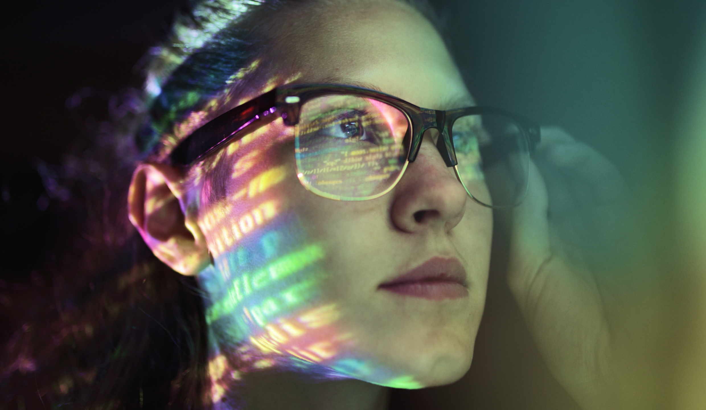 Close-up of a woman wearing glasses with colorful code reflections on her face and lens, looking thoughtfully to the side.