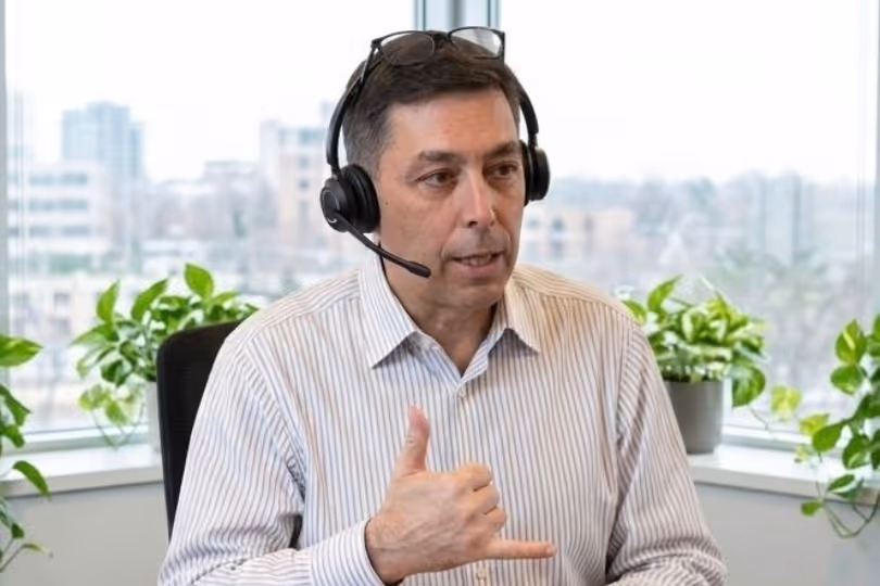 Man wearing headset with microphone, gesturing with thumb and pinky finger in office setting with city view.