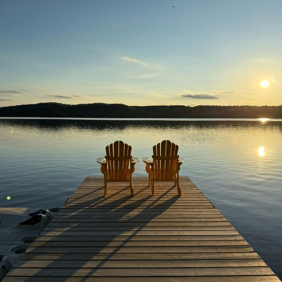 Two empty wooden chairs on a dock facing a calm lake at sunset with hills in the background.