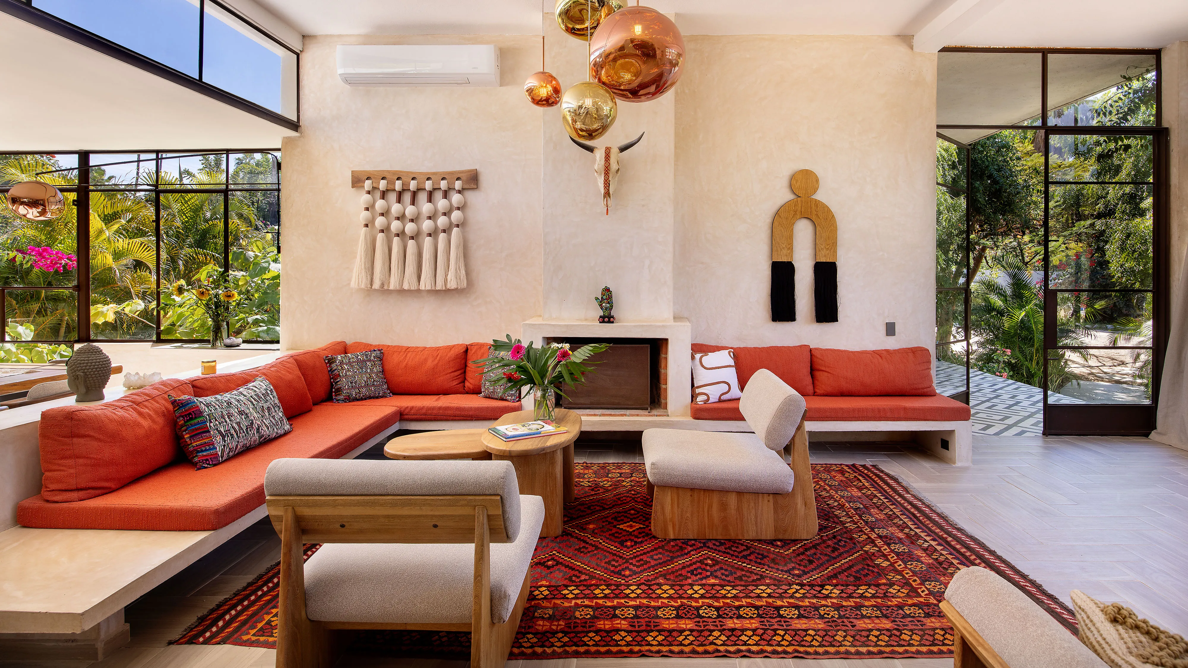 Modern living room with orange sectional sofa, wooden chairs, patterned rug, and large windows showing greenery.