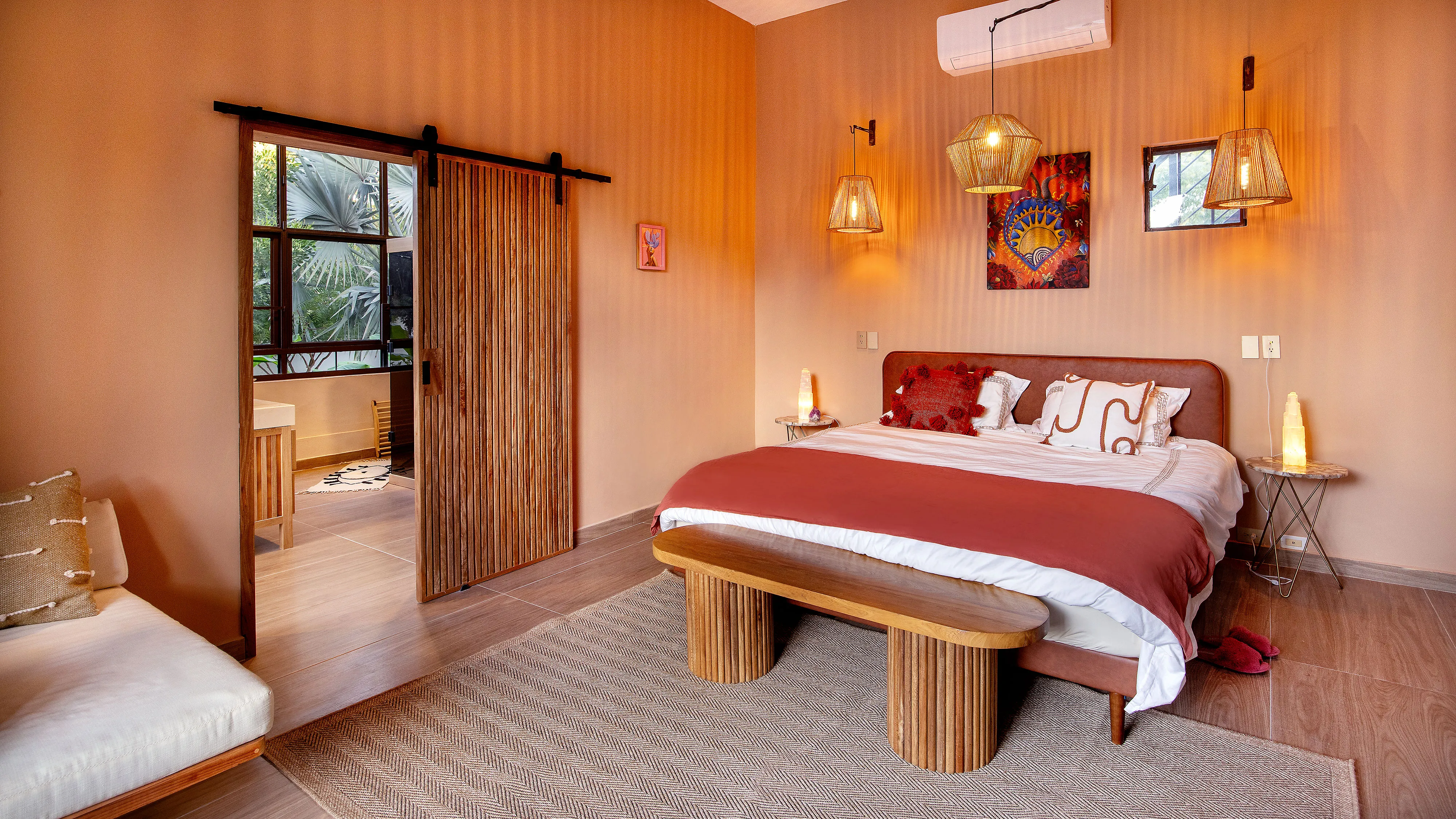 Cozy bedroom with wooden sliding door, bed with red throw, bench, and hanging pendant lamps.