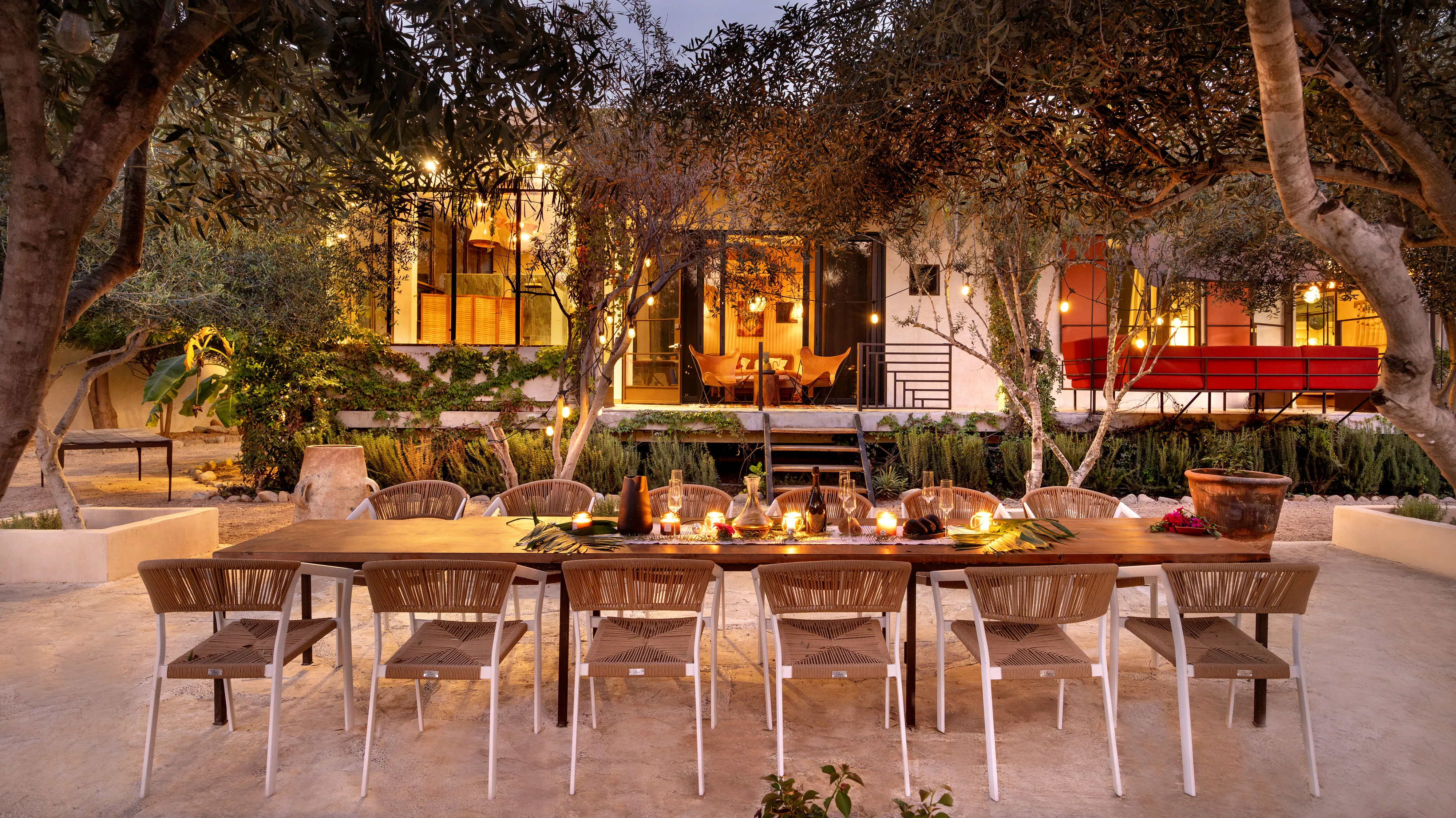 Outdoor dining table with candles and chairs under trees in front of a warmly lit modern house.