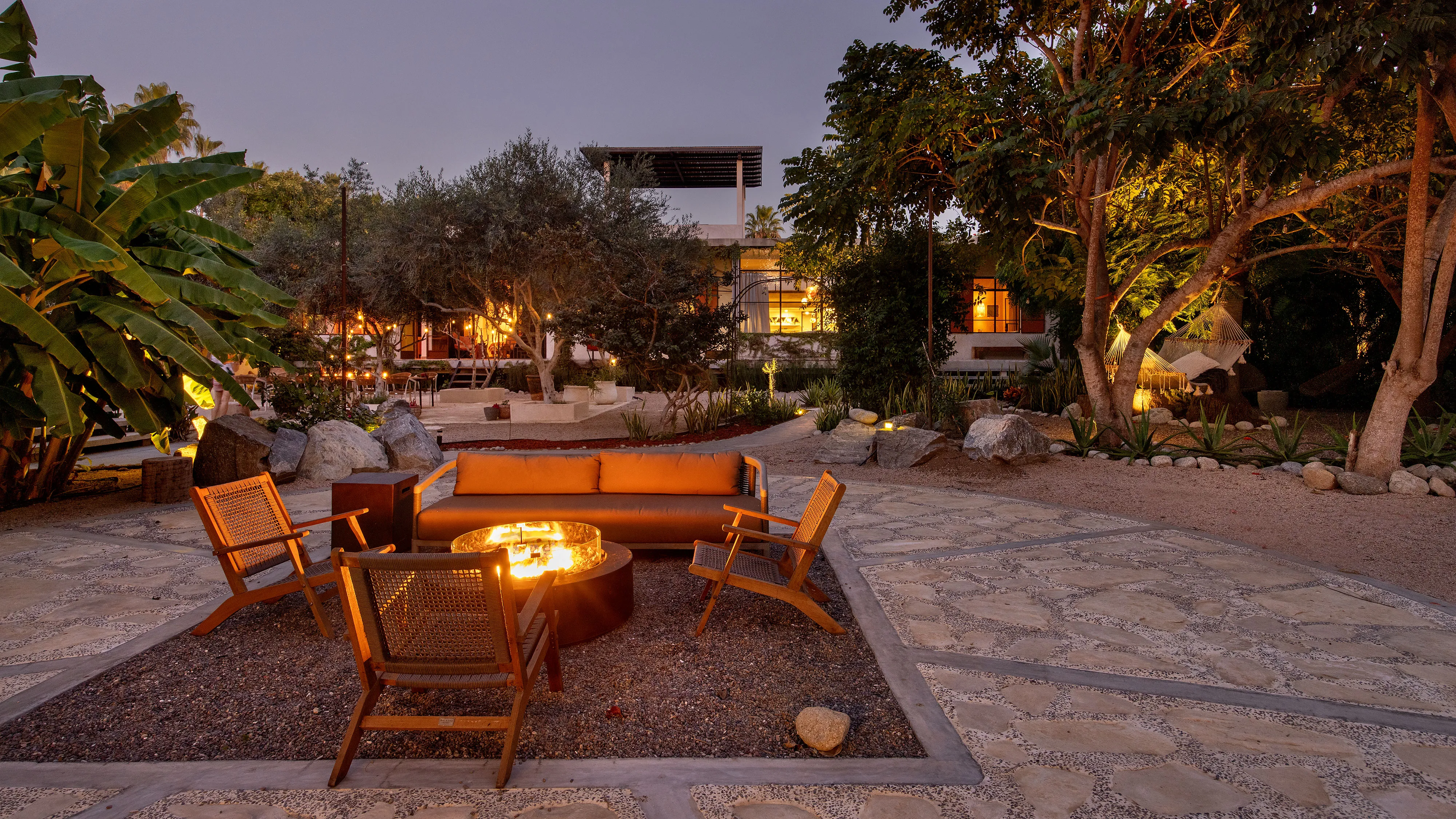Outdoor seating with wooden chairs and sofa around a lit fire pit in a garden at dusk.