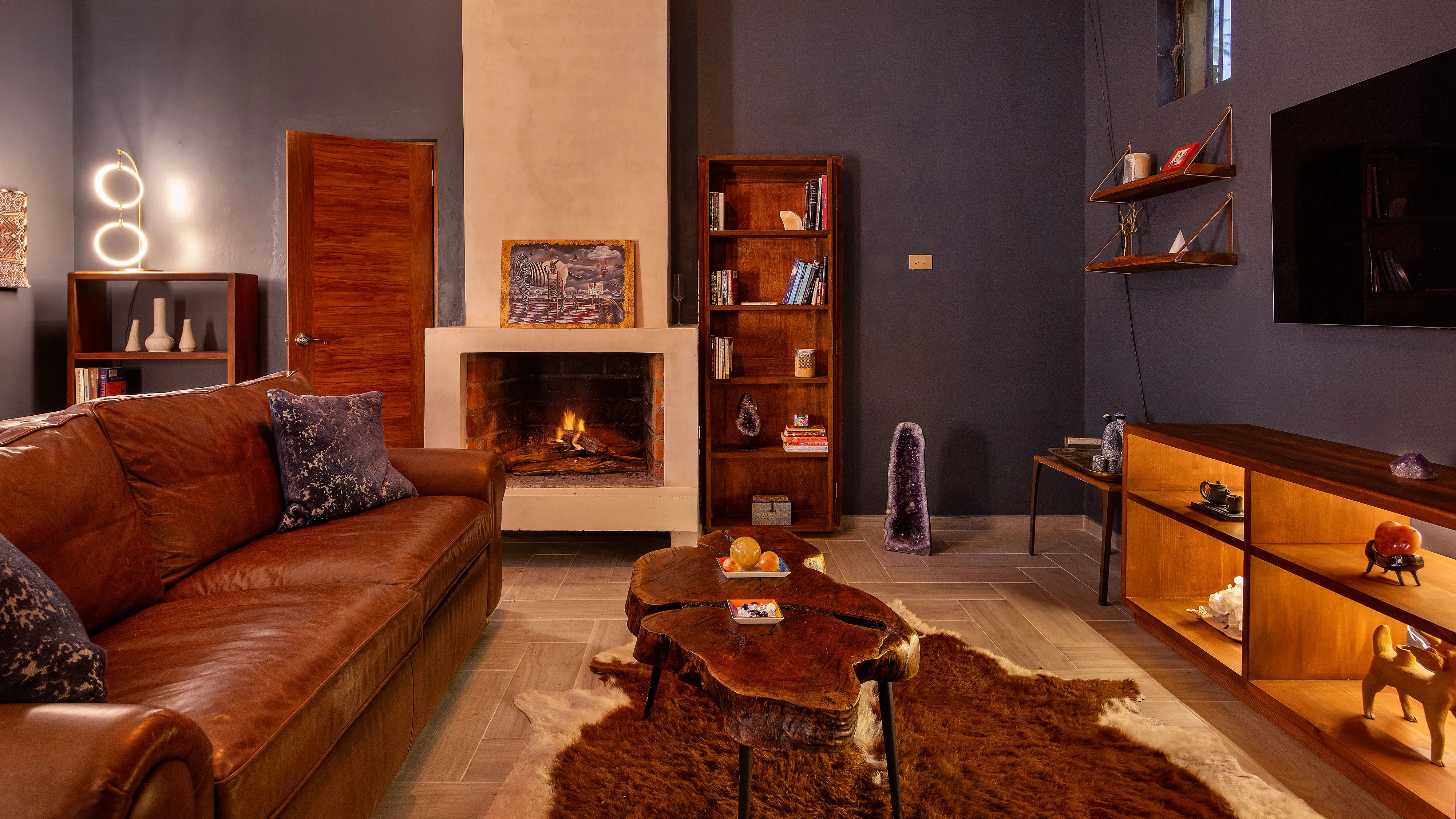 Cozy living room with a brown leather couch, fireplace, wooden shelves, and a rustic coffee table on a rug.