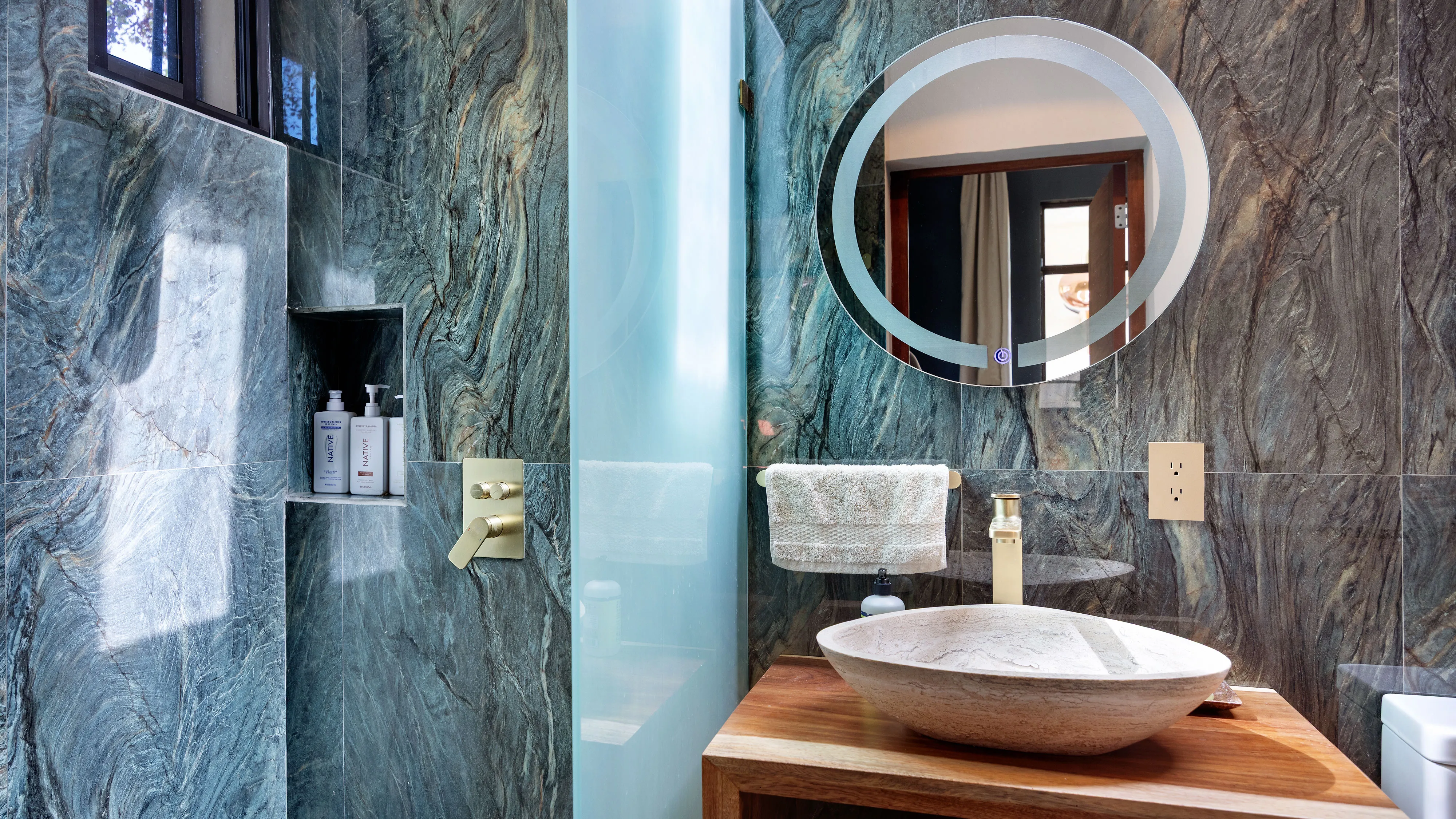 Modern bathroom with green marble walls, round mirror, stone sink, wooden vanity, and frosted glass shower door.
