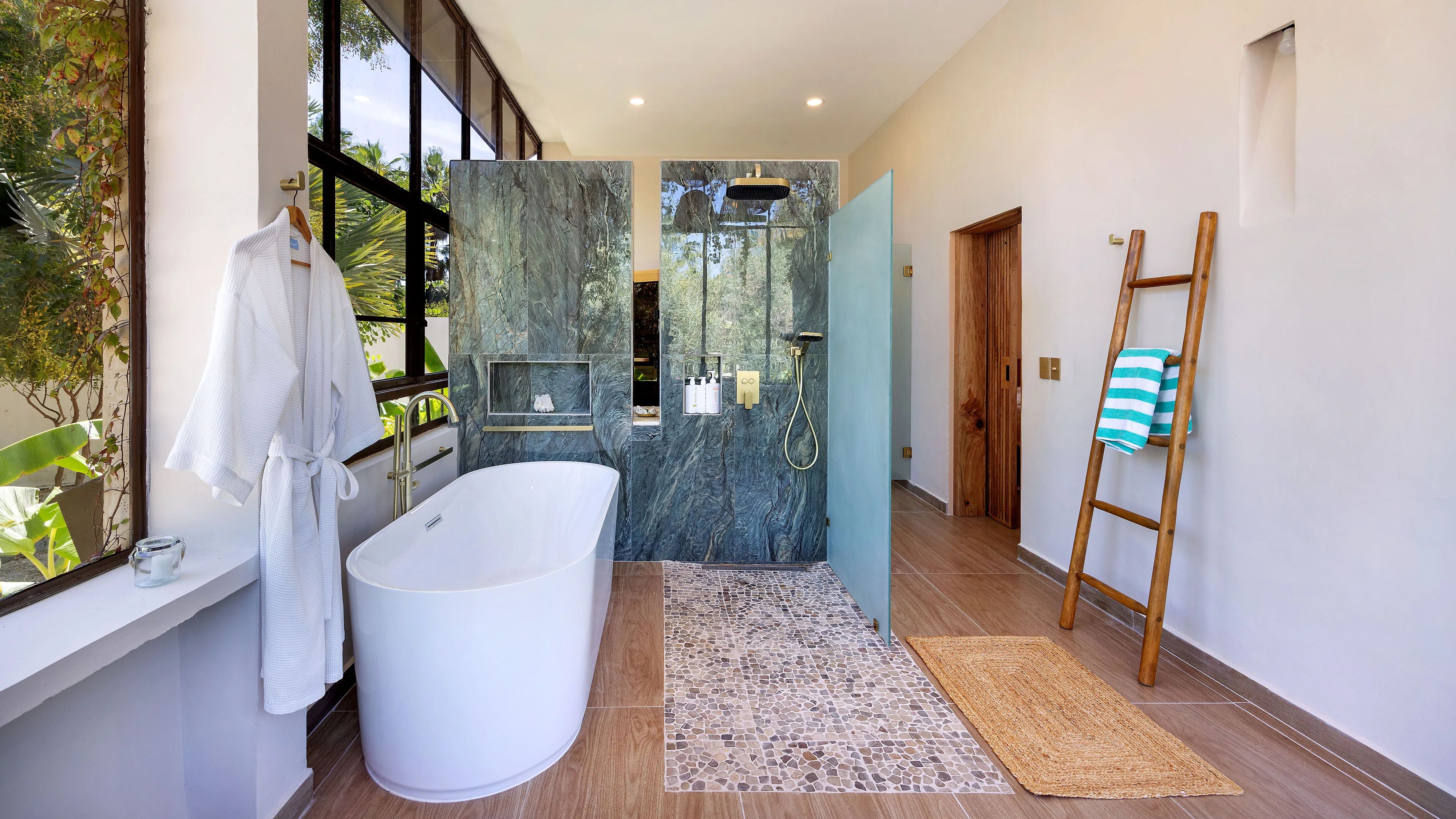 Modern bathroom with freestanding tub, glass shower with marble wall, wooden ladder and large windows.