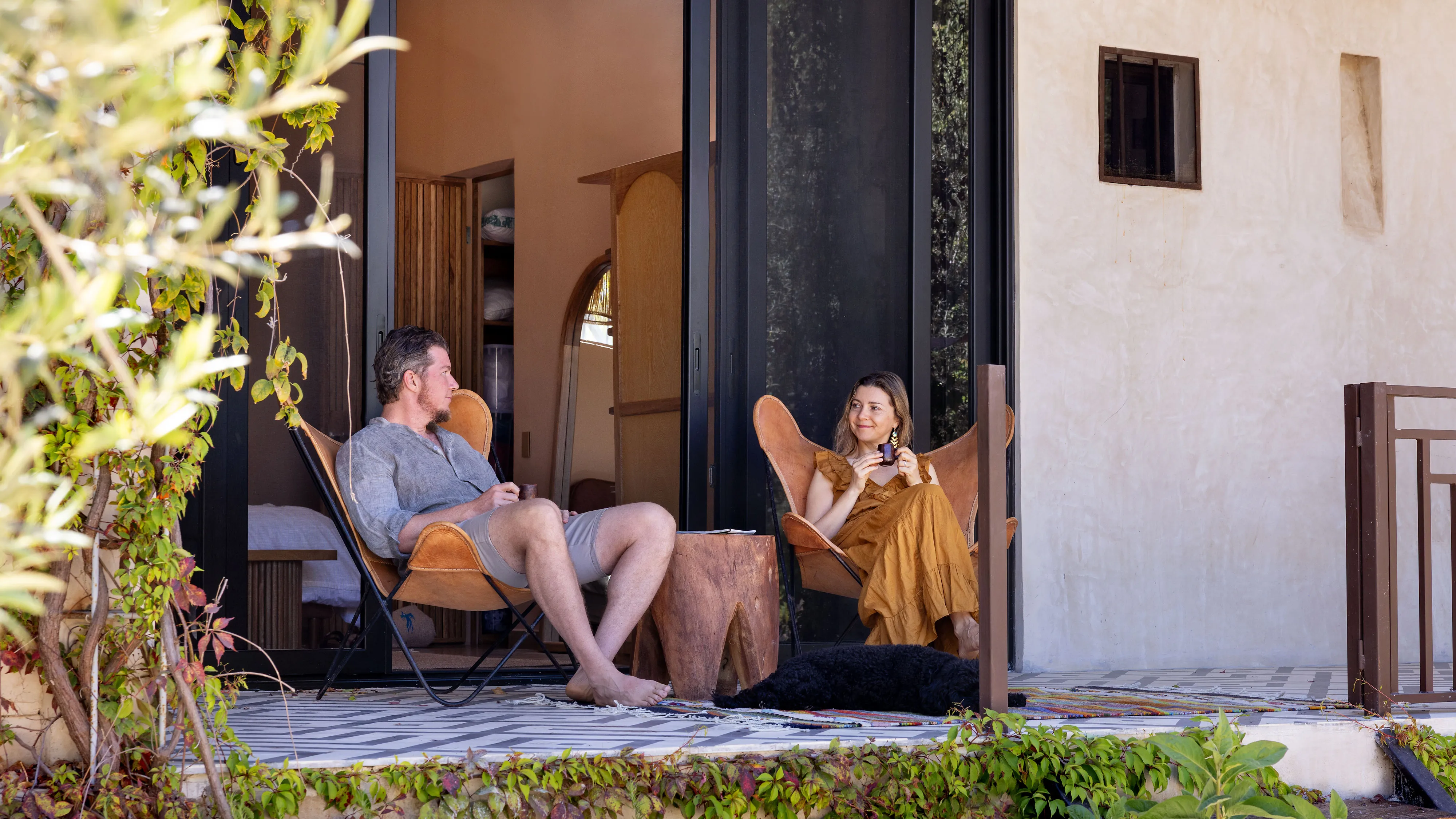 A man and woman sitting in outdoor chairs on a patio, holding coffee cups, with a black dog lying nearby.
