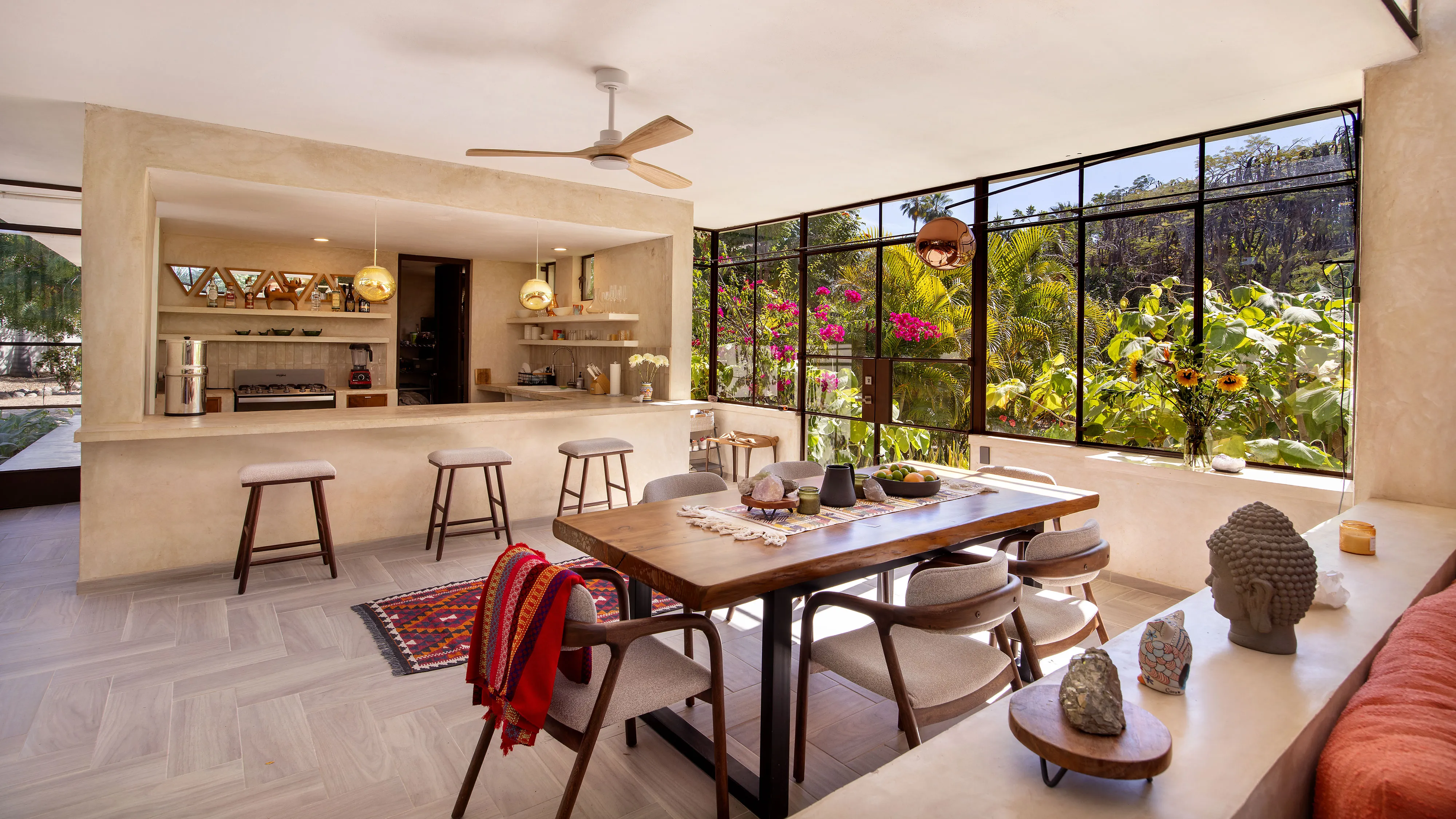 Bright dining area with wooden table, chairs, kitchen bar stools, large windows showing garden outside.