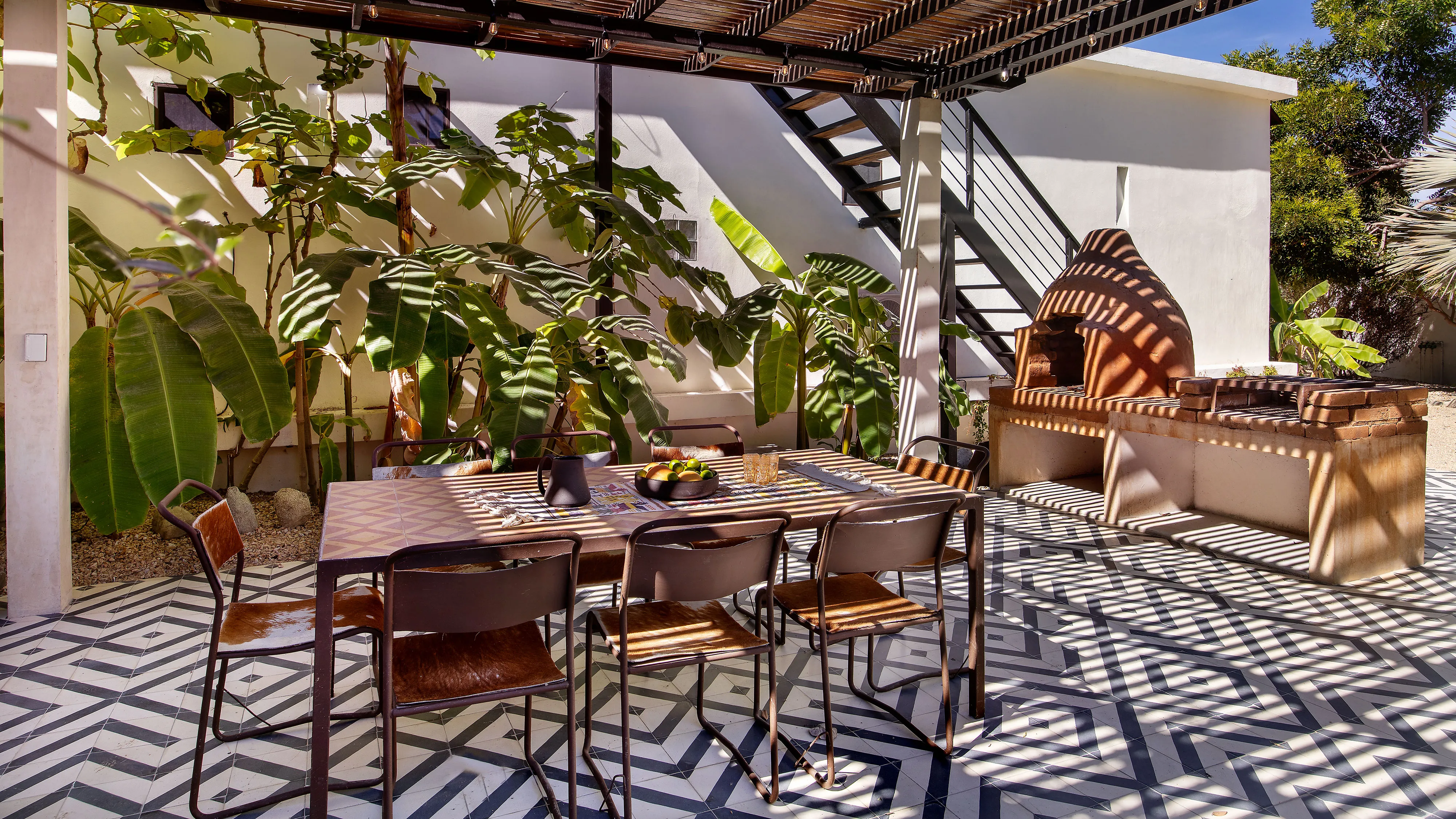 Outdoor dining area with table, chairs, leafy plants, patterned floor, pergola, and brick barbecue oven.