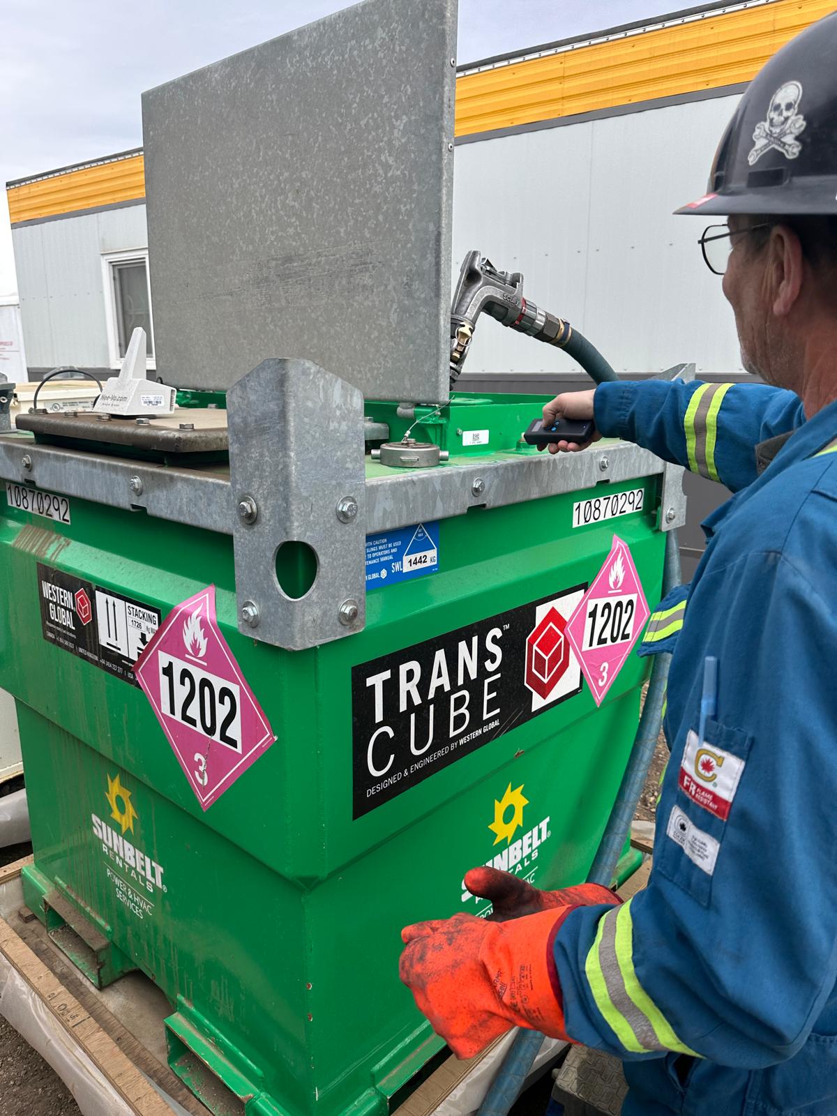 Worker wearing blue coveralls and orange gloves operates a green TransCube fuel container with hazard labels and a fuel nozzle.