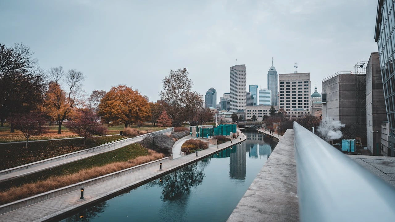 City riverwalk with autumn trees and modern buildings under a cloudy sky.