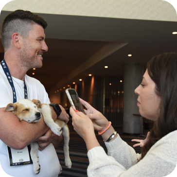 Man holding a sleeping puppy while a woman takes a photo with her smartphone indoors.