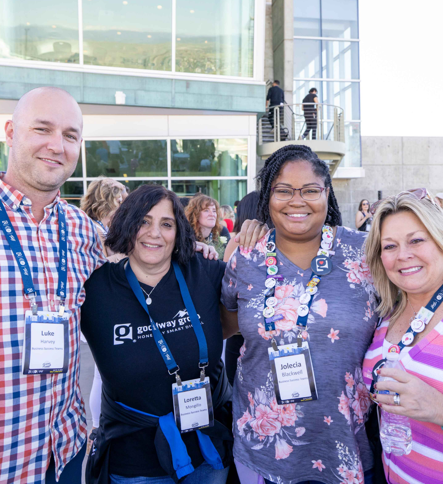 Four smiling adults wearing conference name badges posing together outdoors in front of a building with large windows.