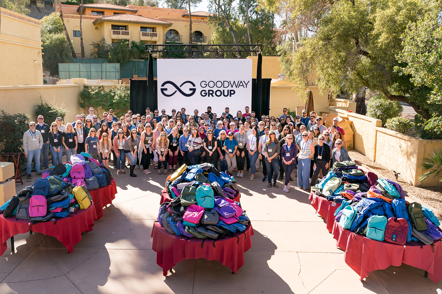 Large group of people standing outdoors in front of a Goodway Group sign with tables of colorful backpacks in front of them.