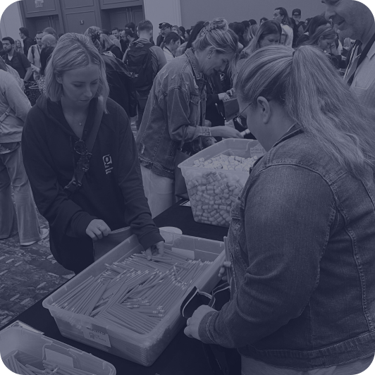 Two women sorting and packing supplies in large plastic bins at a crowded indoor volunteering event.
