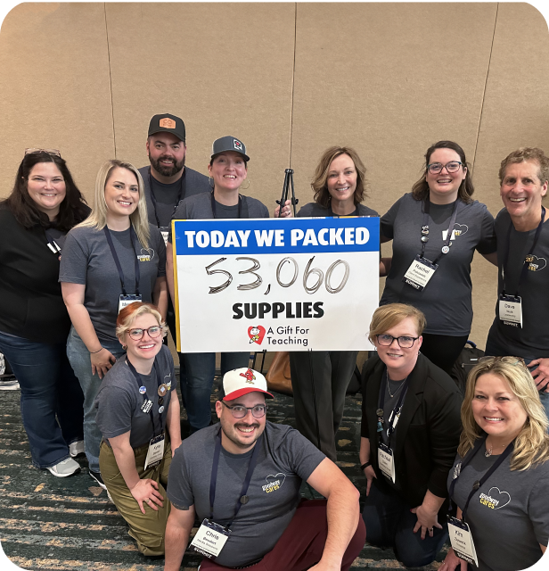 Group of eleven smiling people wearing matching gray shirts holding a sign that reads, 'TODAY WE PACKED 53,060 SUPPLIES A Gift For Teaching.'
