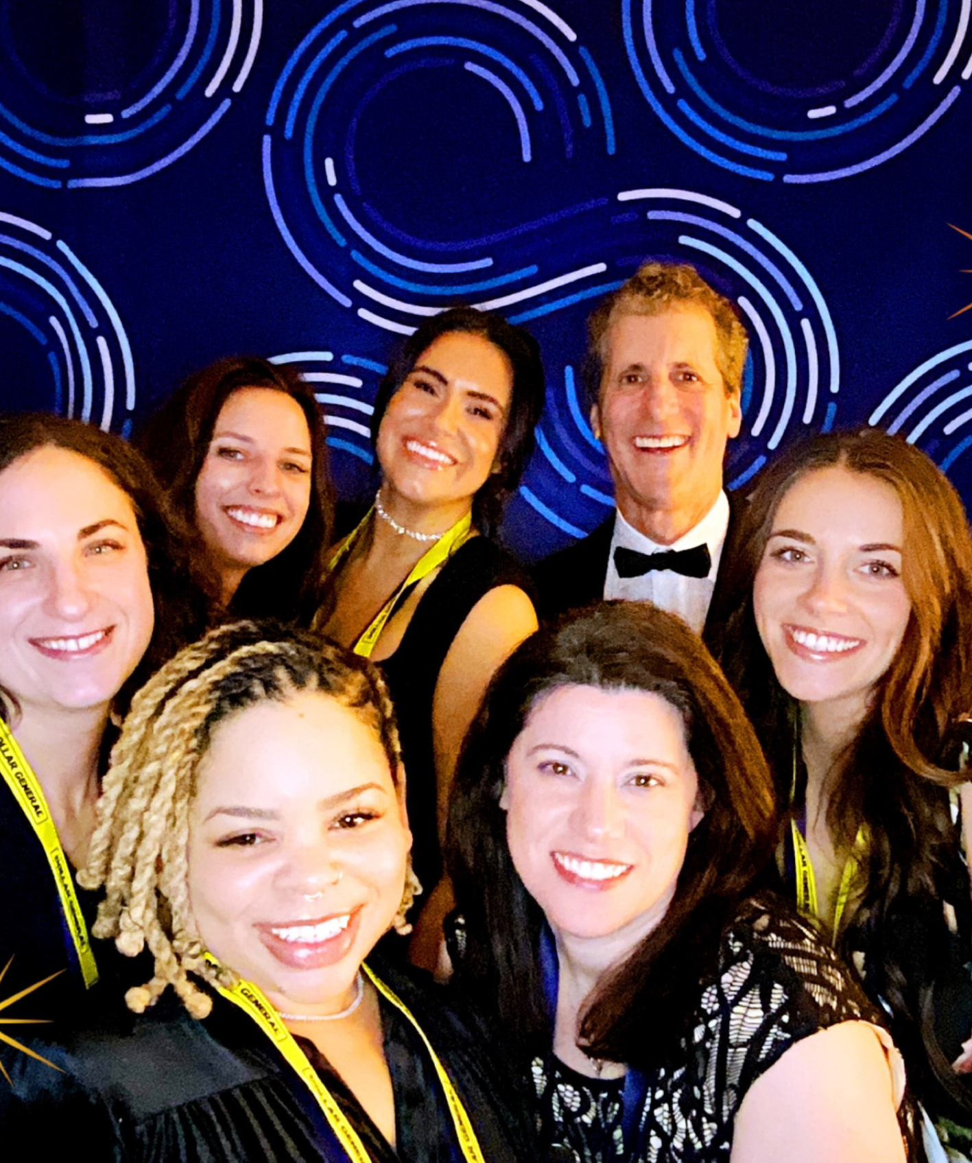 Group of six women and one man smiling in front of a blue backdrop with circular patterns, several wearing yellow lanyards.