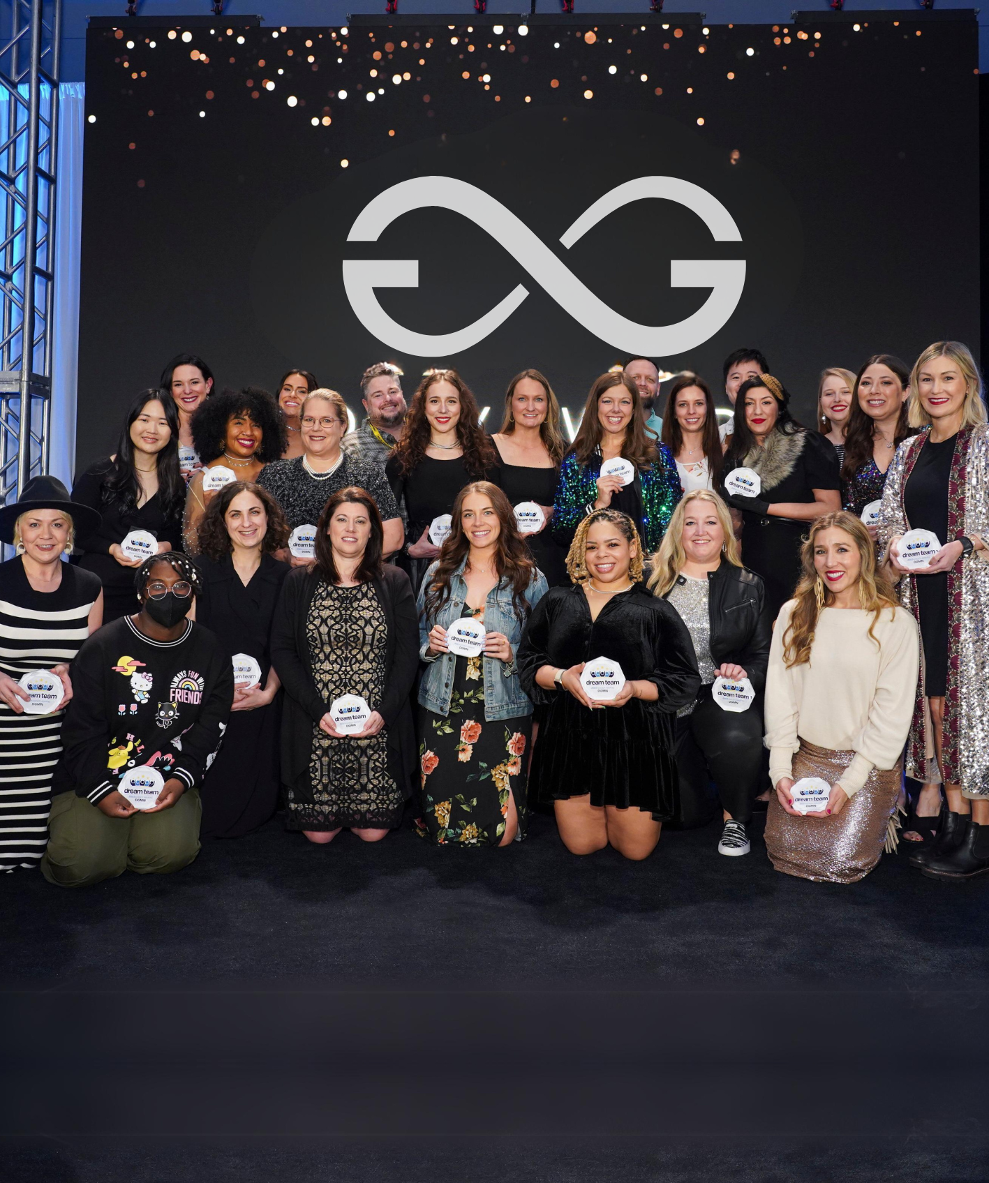 Group of diverse people posing on stage holding 'dream team' plaques beneath a large infinity logo.