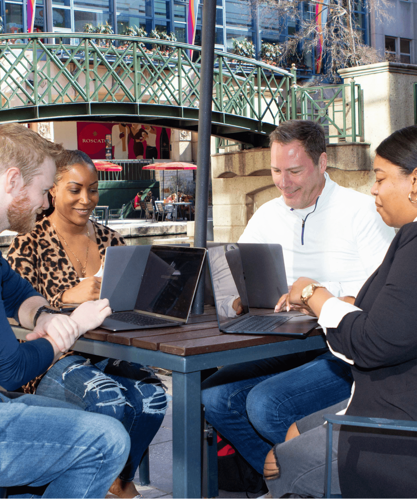 Four diverse adults gathered around a table outdoors, each using a laptop and engaged in discussion.