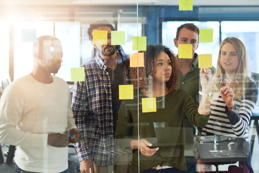 A diverse group of five colleagues collaborating and placing sticky notes on a glass wall in a modern office.