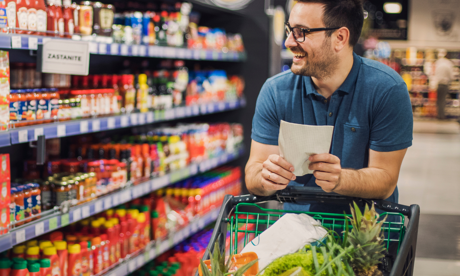 Man wearing glasses smiling while holding a shopping list with a grocery cart filled with fruits and vegetables in a store aisle.