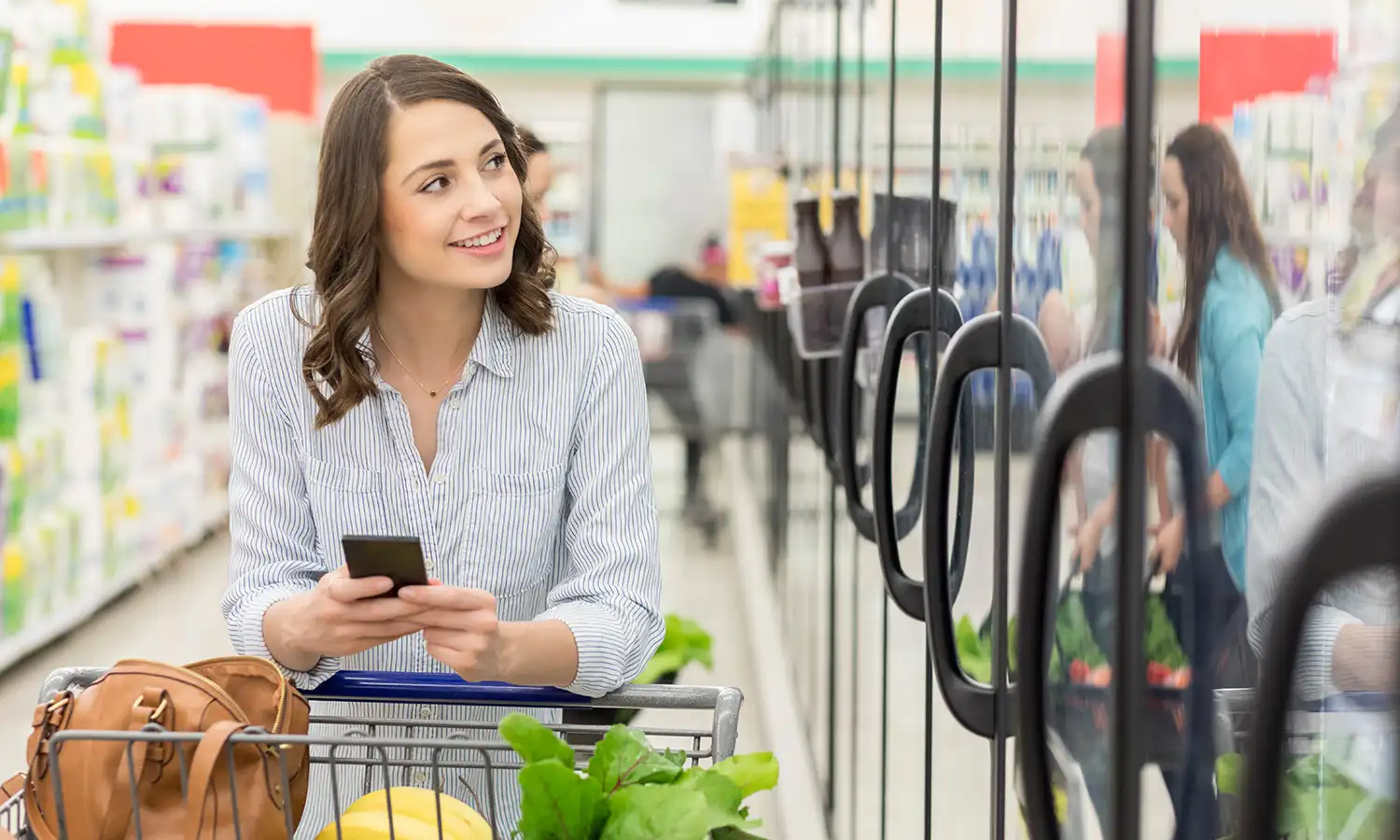 Smiling woman holding a smartphone and standing behind a grocery cart filled with produce in a supermarket aisle.