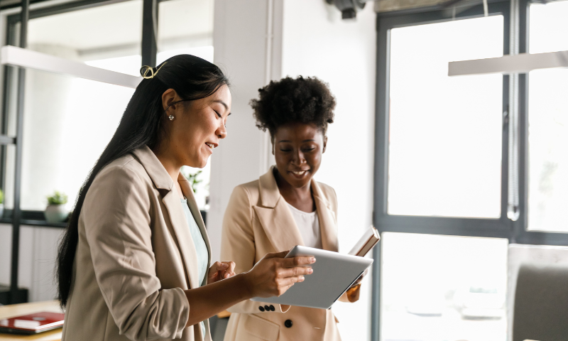 Two professional women in beige blazers discussing content on a tablet in a bright office.
