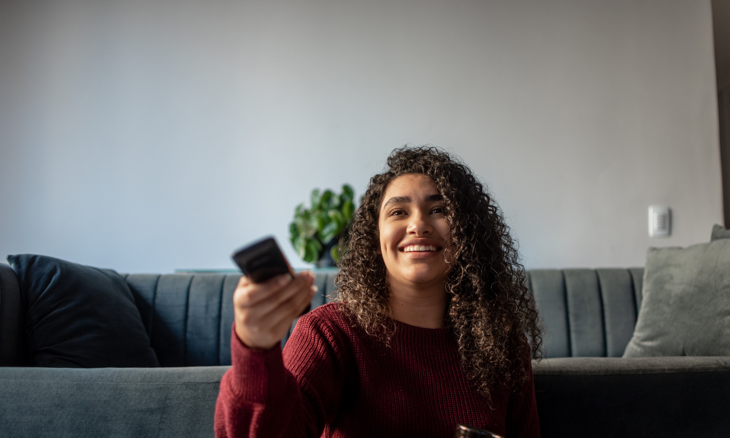 Woman with curly hair sitting on a couch, smiling and holding a TV remote control.