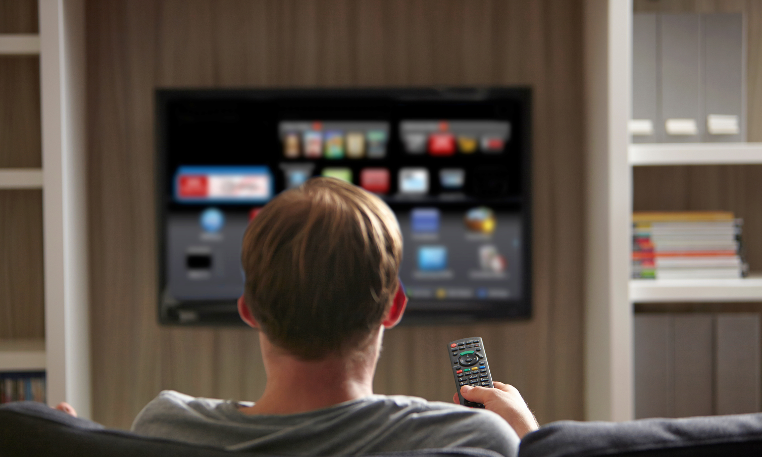 Man sitting on a couch holding a remote control, watching a smart TV with app icons on the screen.