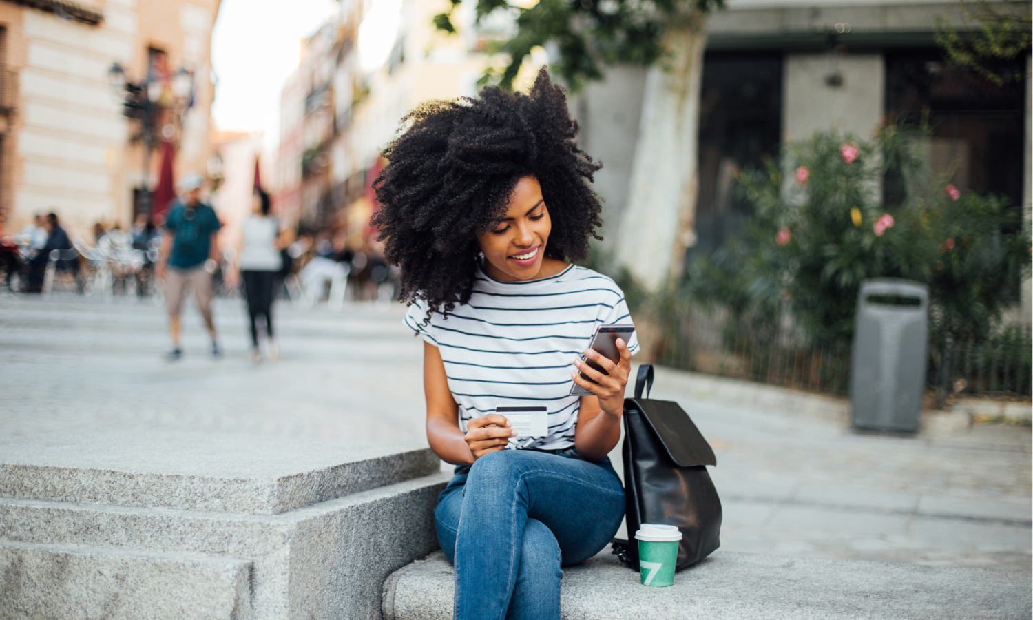 Smiling woman sitting on stone steps outdoors, holding a credit card and looking at her smartphone with a black backpack and coffee cup beside her.