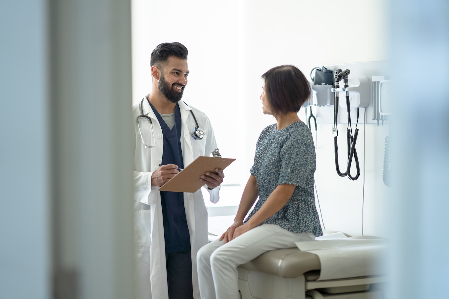 Male doctor in a white coat smiling and holding a clipboard while talking to a female patient sitting on an exam table in a medical office.