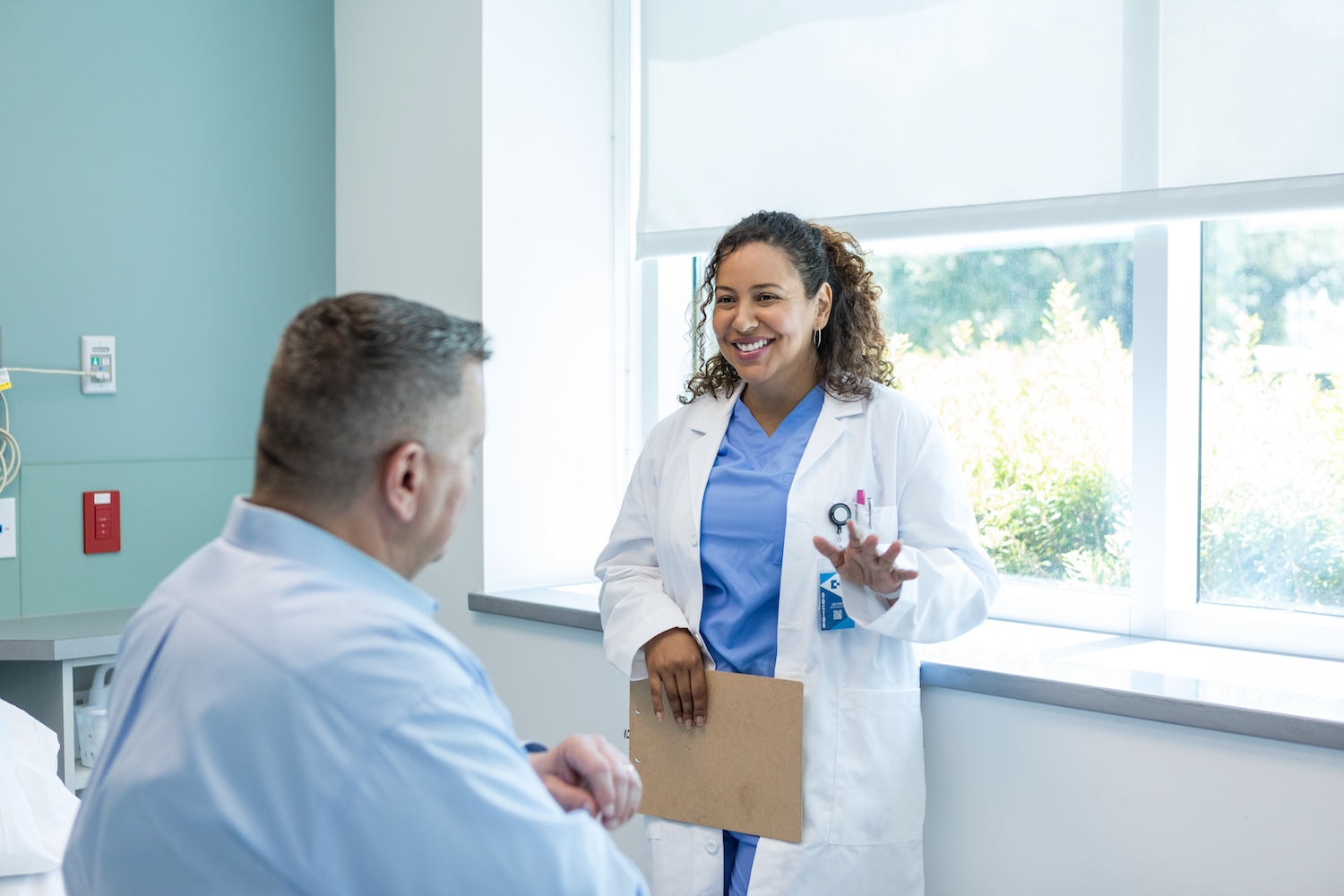 Female doctor in blue scrubs and white coat holding a clipboard, smiling and talking to a seated male patient in a hospital room.