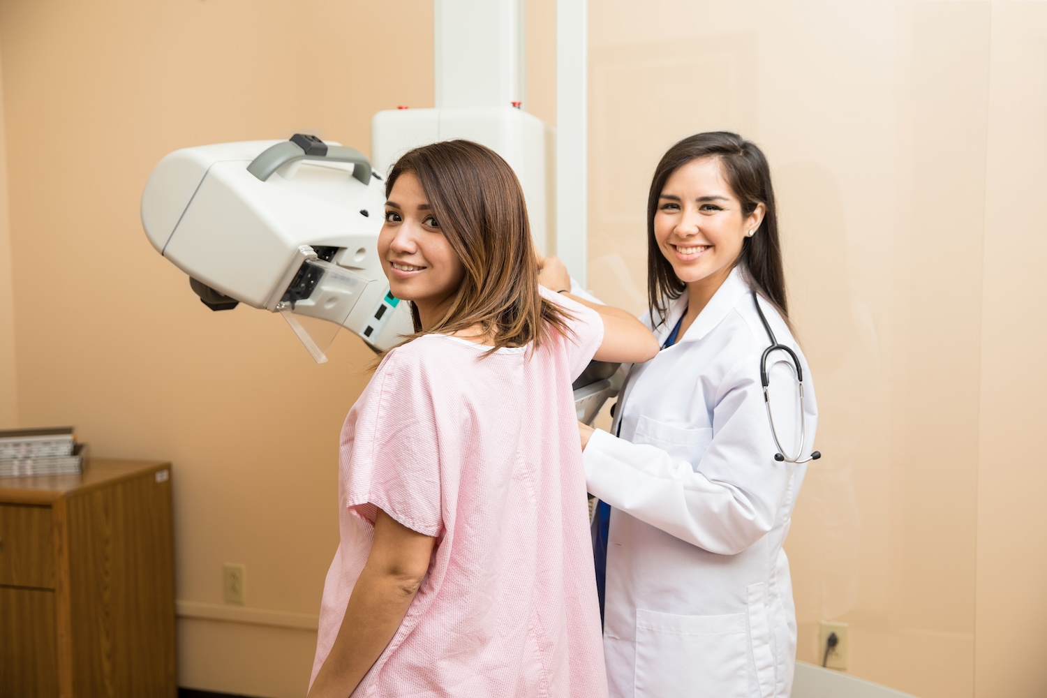 Woman in pink hospital gown undergoing a mammogram performed by a female healthcare professional with a stethoscope.