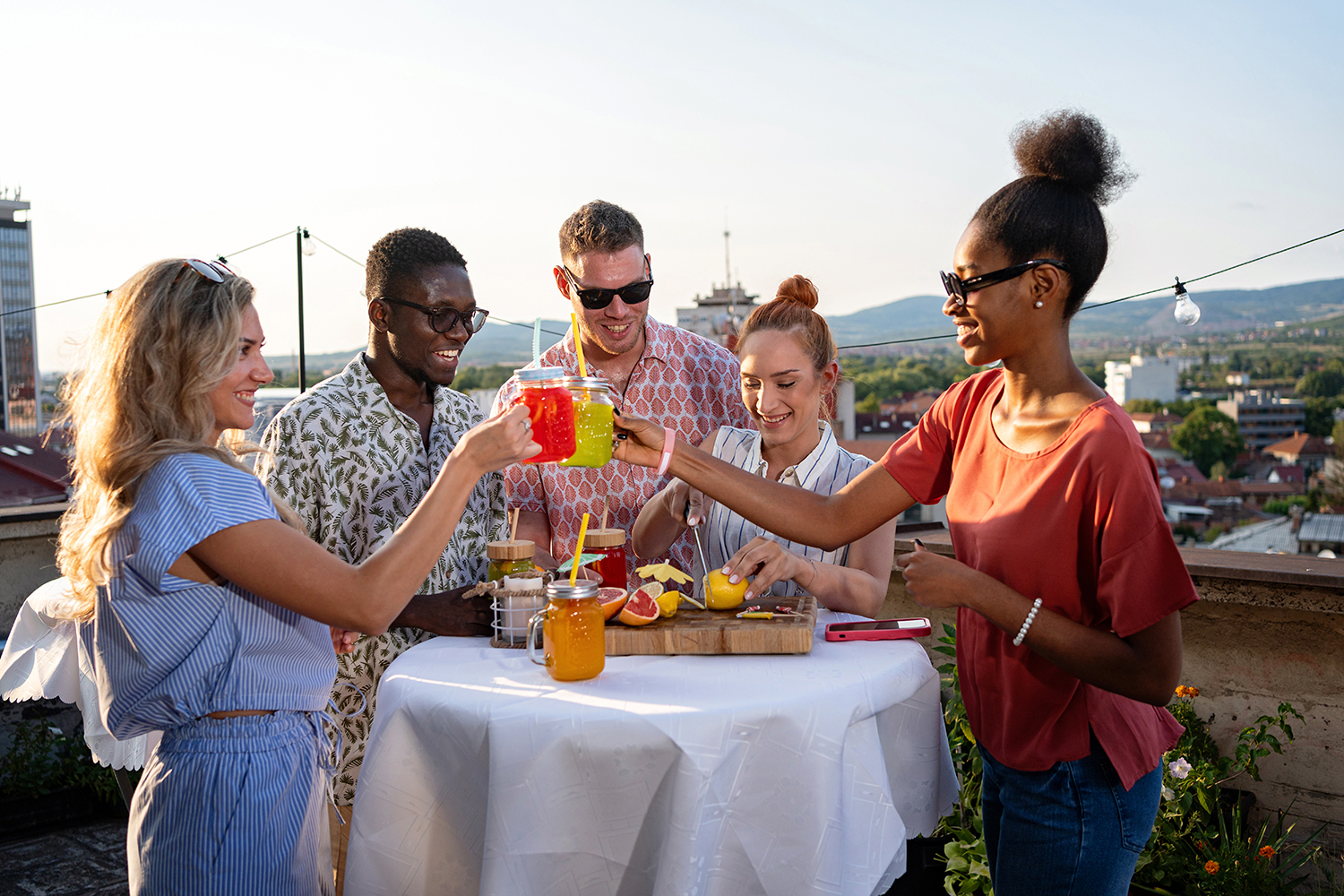 Five friends on a rooftop toasting with colorful drinks around a table with fruit and jars.