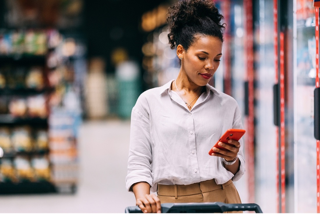 Woman in a white shirt looking at her phone while pushing a shopping cart in a grocery store aisle.