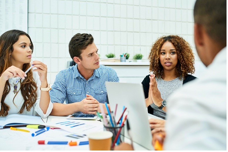 Group of four young professionals having a discussion around a table with papers, pens, and a laptop in a modern office.