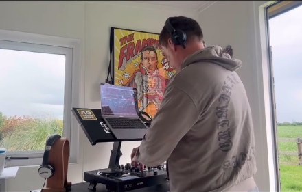 Man wearing a gray hoodie and headphones DJing indoors near a window with a laptop and mixer setup.
