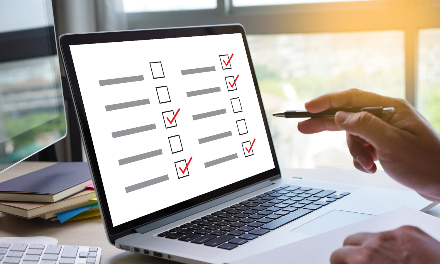 Laptop screen displaying a checklist with several checkboxes marked with red checkmarks, and a hand holding a pen pointing at the screen.